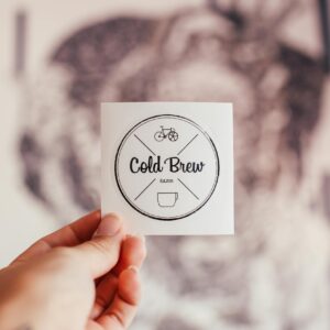 Unrecognizable person demonstrating coffee shop badge with Cold Brew inscription and creative design in hand while standing on blurred background